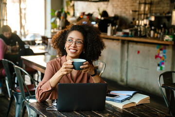 Young african american woman using laptop and drinking coffee in cafe - Powered by Adobe