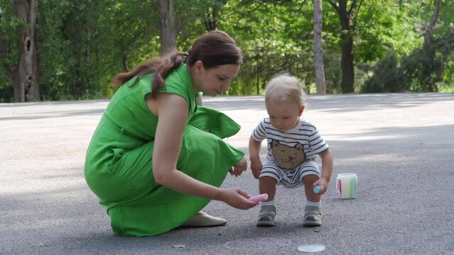 Mother and little child having fun drawing on asphalt in summer park, toddler boy playing with colored sidewalk drawing chalk on a path on sunny day. Outdoor playground activity for children