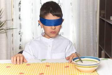 An 8-year-old Caucasian boy sitting at the table at home is blindfolded with a serious gesture. The...
