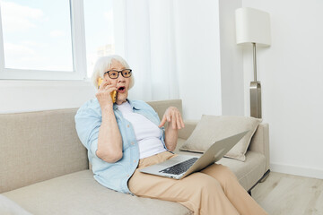 a cheerful, pleasant woman is sitting on a sofa in a bright room and working at a laptop on her lap happily talking on the phone