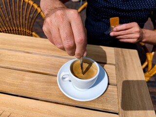 Fototapeta na wymiar White caucasian male hand stirs a freshly brewed cup of coffee on a patio terrace on a wooden table. There are no recognizable people or trademarks in the shot.