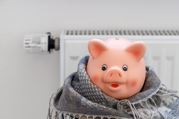 Pink piggy bank with warm scarf on light background with heating radiator. Expensive and rising heating costs and savings energy concept