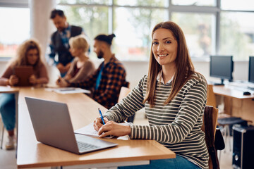 Happy mid adult student using laptop while writing notes in classroom and looking at camera.