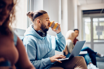 Young student drinks coffee while e-learning on laptop in hallway.