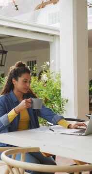 Vertical video of happy biracial woman drinking coffee and using laptop
