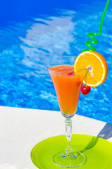 Orange fresh with a red cherry and an orange inside on a glass, against the backdrop of a blue pool.