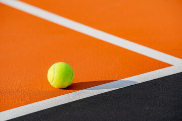 Yellow tennis ball at orange tennis court with white baseline and black out of bounds	