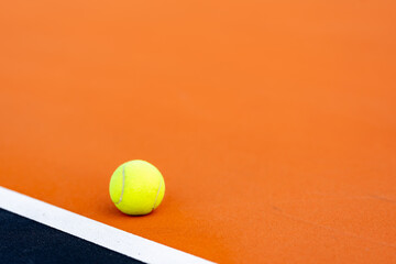 Yellow tennis ball at orange tennis court with white baseline and black out of bounds	