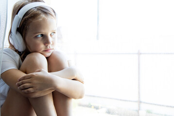 Portrait of a little girl sitting on the windowsill and thoughtfully looking out the window. Hugs his knees and is sad listening to music with headphones.