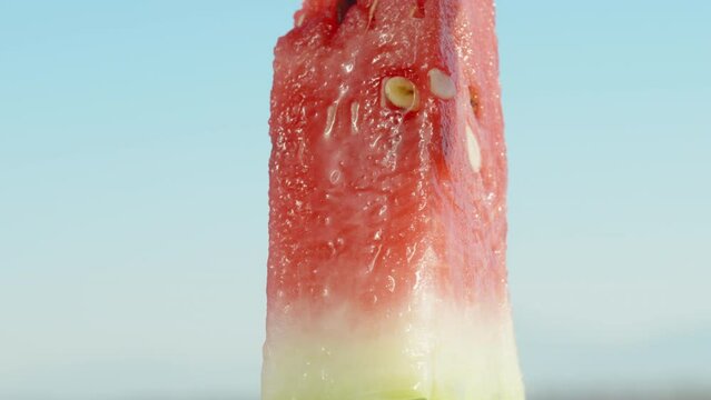 A piece of red watermelon rotating against the blue sky. The juice flows down it. Close-up.