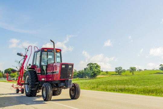 An International 5088 tractor pulling a tedder down a country road on a summer day with a hay field in the background.