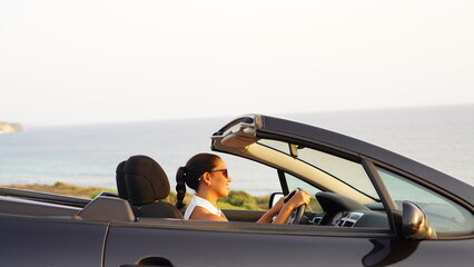 Woman alone in a black convertible car looks at the sunset in front of the sea and is moved by how...