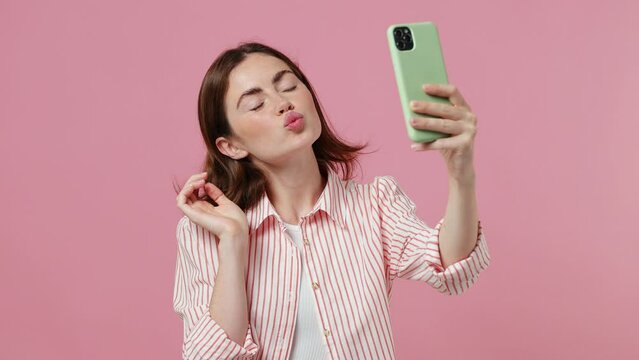 Young cheerful woman she wearing shirt t-shirt doing selfie shot on mobile phone post photo on social network isolated on plain pastel light pink background studio portrait. People lifestyle concept