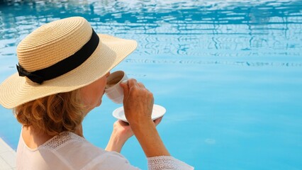 Close up of a woman aged 50-55 in a straw hat drinking from a cup of coffee next to a blue swimming...