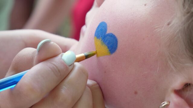 Drawing on unrecognizable child's cheek a heart in the yellow-blue colors of the Ukrainian flag. Peace concept, protest against war