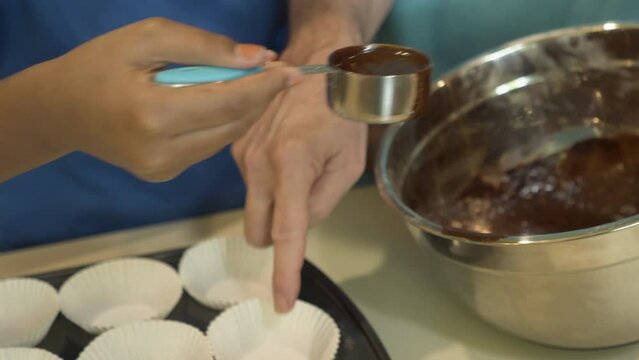 A senior adult helping a child pour a chocolate batter into a prepared cupcake tin with paper liners.