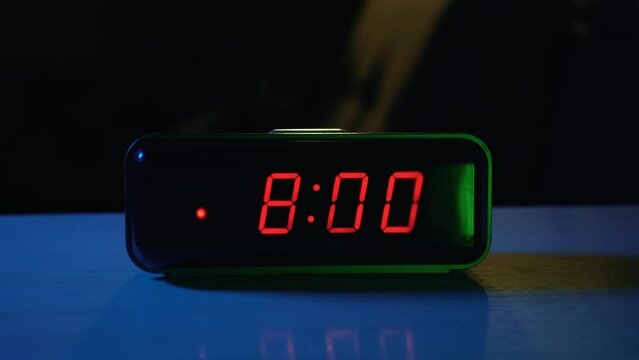 The electronic alarm clock on the table rings at 8:00 in the morning. The hand turns it off by pressing a button. Start of the day and wake up for work or study