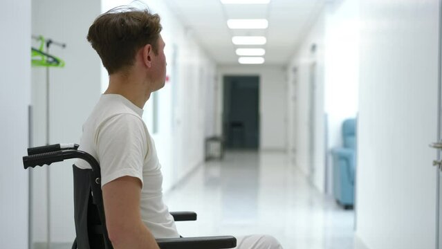 Side view devastated Caucasian ill man in wheelchair in hospital corridor looking away waiting and looking at camera. Portrait of sad depressed patient in medical clinic alone. Disability frustration