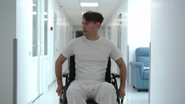 Man rolling wheelchair along hospital corridor looking around. Front view portrait of Caucasian male patient looking for doctor in medical clinic. Disability and medicine concept