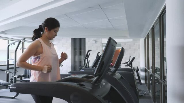 Side view, medium shot of long hair Asian middle-aged woman, running on treadmill, workout alone at indoor fitness gym. Sport, health, wellbeing, weight loss concept.