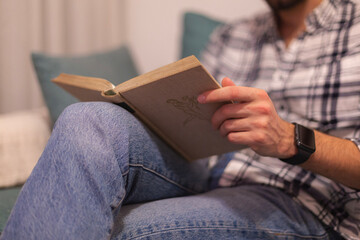 Closeup view of male hands holding book. Young man relaxing at home sitting in a couch. Selective focus on hand, blurred background. Horizontal.
