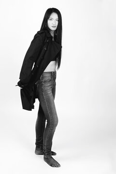 Beauty, fashion and style concept. Beautiful Asian woman with long dark hair studio portrait in bright background with copy space. Model wearing jeans and black trench coat. Black and white image