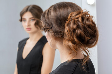 Fototapeta premium A young attractive woman with beautiful makeup in a black dress straightens her hair in front of a mirror.