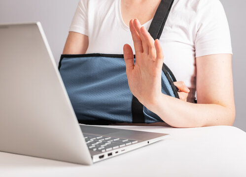 Woman with arm in sling using laptop for video call. Female waving hand to greet smb while talking online. Work, study from home due to injury. High quality photo