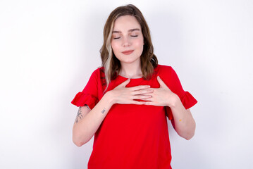 young caucasian woman wearing red T-shirt over white background  smiling with hands on chest with closed eyes and grateful gesture on face. Health concept.