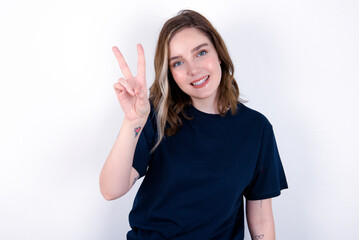 Obraz na płótnie Canvas young caucasian woman wearing black T-shirt over white background smiling and looking happy, carefree and positive, gesturing victory or peace with one hand