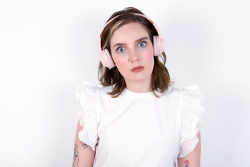 Fototapeta premium Serious displeased young caucasian woman wearing white T-shirt over white background looks puzzled at camera being angry wears stereo headphones listens music while walking at street
