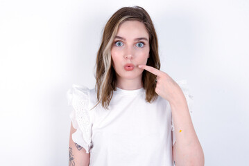 Charming young caucasian woman wearing white T-shirt over white background pointing on pout lips with forefinger, showing effect after lifting procedure,
