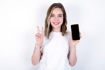 young caucasian woman wearing white T-shirt over white background holding modern device showing...