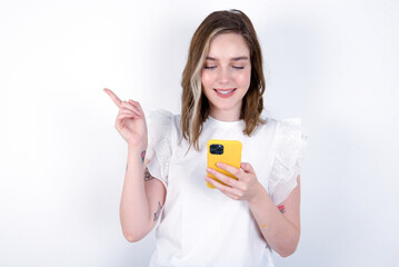 Smiling young caucasian woman wearing white T-shirt over white background pointing finger at blank space holding phone in one hand