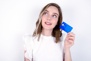 Lovely young caucasian woman wearing white T-shirt over white background showing credit card and looking away at copy space. Pensive