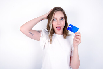 Impressed funny young caucasian woman wearing white T-shirt over white background hand on head holding bank card
