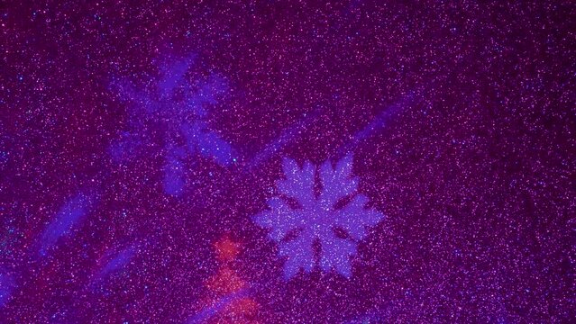 A carousel of light beams of different colors is set against a constantly flashing background that changes colors. Flying colored silhouettes of Christmas trees and snowflakes.