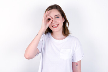 young caucasian woman wearing white T-shirt over white background with happy face smiling doing ok sign with hand on eye looking through finger.