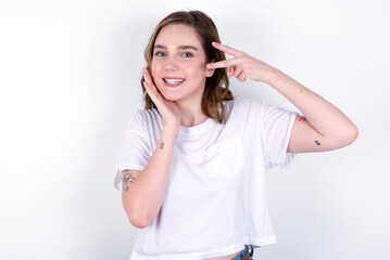 young caucasian woman wearing white T-shirt over white background making v-sign near eyes. Leisure lifestyle people person celebrate flirt coquettish concept.