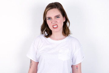 Fototapeta premium Mad crazy young caucasian woman wearing white T-shirt over white background clenches teeth angrily, being annoyed with coming noise. Negative feeling concept.