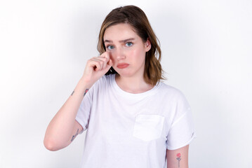 Unhappy young caucasian woman wearing white T-shirt over white background crying while posing at camera whipping tears with hand.