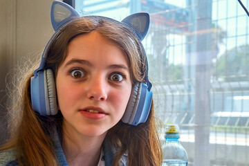 Horizontal shot of a happy cute beautiful natural teenage girl wearing a denim jacket and blue headphones, she is riding a train or bus, sitting near the window and looks surprised. Positive emotions