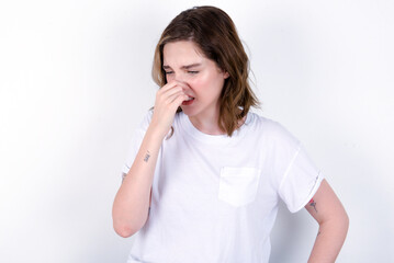 young caucasian woman wearing white T-shirt over white background , holding his nose because of a bad smell.