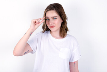 Upset young caucasian woman wearing white T-shirt over white background shapes little gesture with...