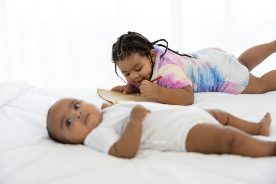 cute African baby lying on bed and her sister writing a book or doing homework at home