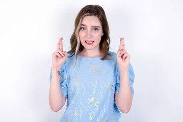young caucasian woman wearing blue T-shirt over white background holding fingers crossed with...
