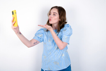 young caucasian woman wearing blue T-shirt over white background blows air kiss at camera of smartphone and takes selfie, sends mwah via online call.