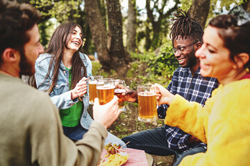 multiracial group of friends gather outside at picnic to chat and have beers together - people holding beer mugs for a celebratory toast - focus on the African man