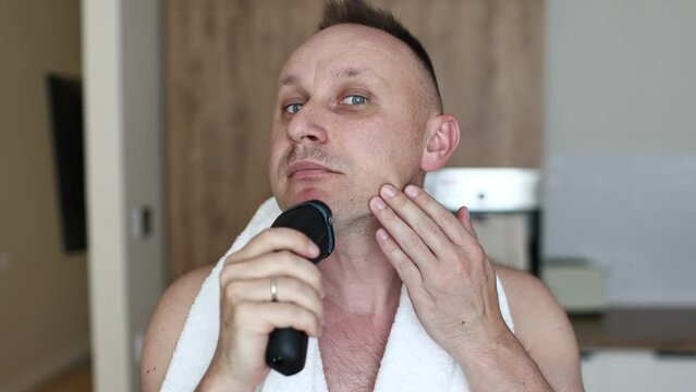 young unshaven man with towel on shoulders shaving with modern electric razor trimer while looking at camera, indoors. Daily routine male self-care. man shaves with an electric razor in the kitchen.