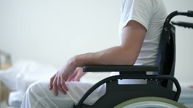 Unrecognizable man in wheelchair waiting for nurse making bed at background. Side view Caucasian ill patient in hospital with doctor helping. Rehabilitology and medicine concept